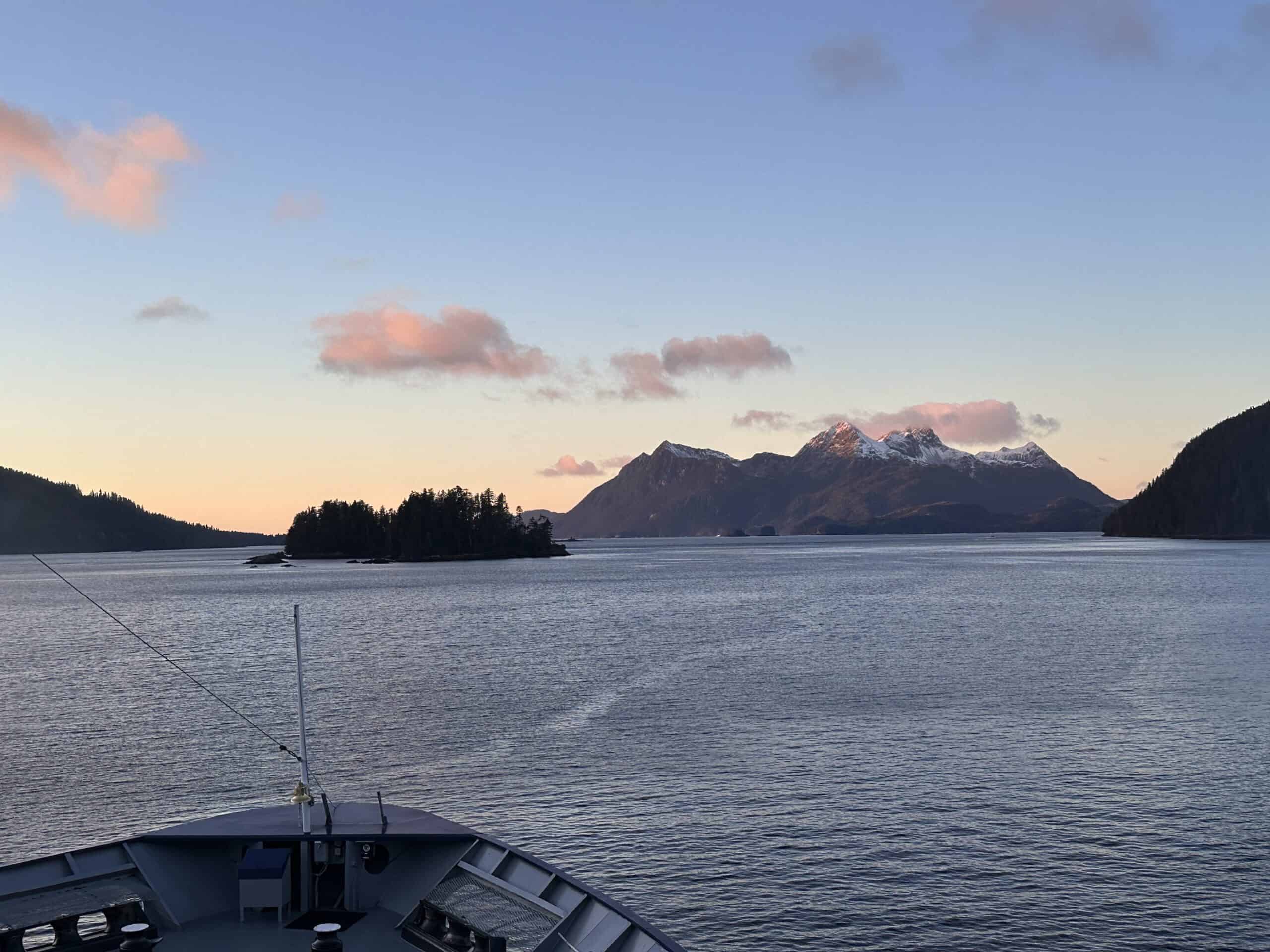 view of an Alaskan sunset behind mountains, standing at the bow of a ship.