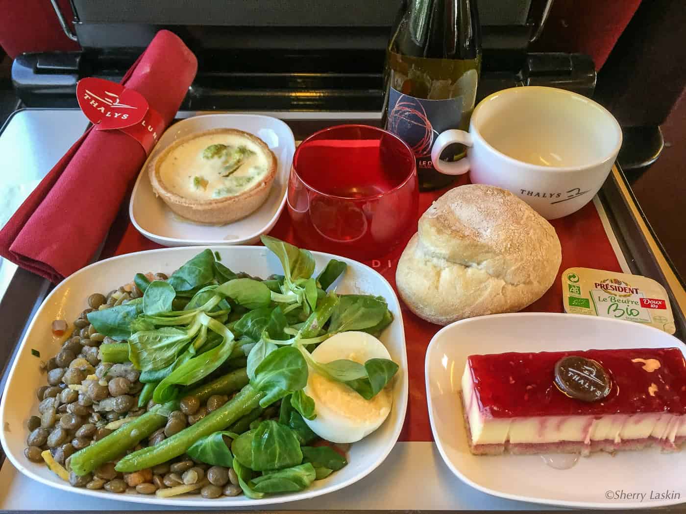 Lunch aboard Thalys train served a lentil, greens and egg salad, bottle of wine, dinner roll with butter and cheesecake.